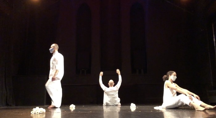 Alternative text: Alethea Pace, Arthur Aviles, and Richard Rivera dance on a black marley floor illuminated by stage lighting. They wear all white garb including a white face covering.